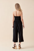 Load image into Gallery viewer, Back view of model wearing the Shiloh Smocked Tube Top Jumpsuit in Black paired with block-heel sandals to complete this chic, warm-weather outfit.