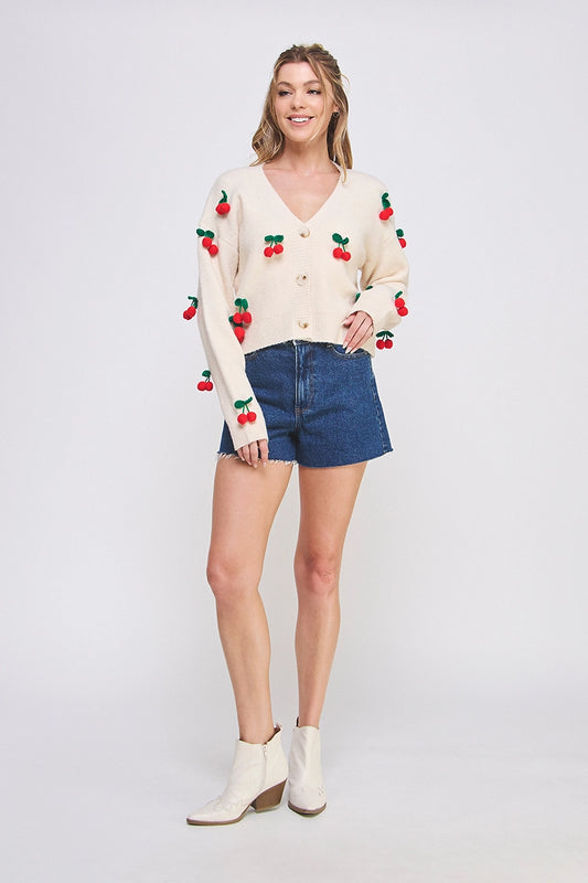 Woman wearing a cream cardigan with red cherry patterns, denim shorts, and white boots on a light gray background